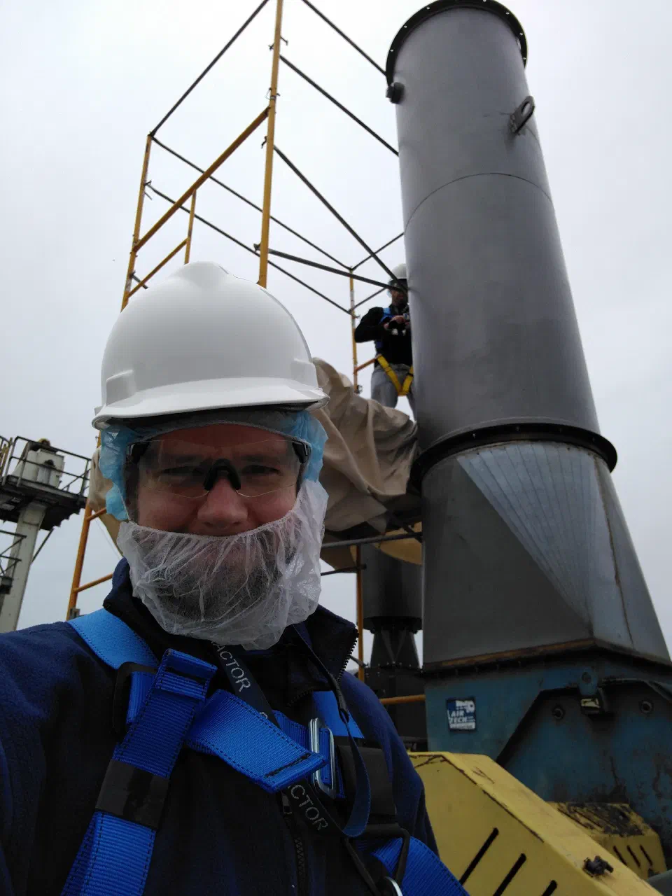 Phil in a hard hat, safety glasses, beard net and fall arrest harness on a roof, beside an industrial chimney being installed from scaffolding